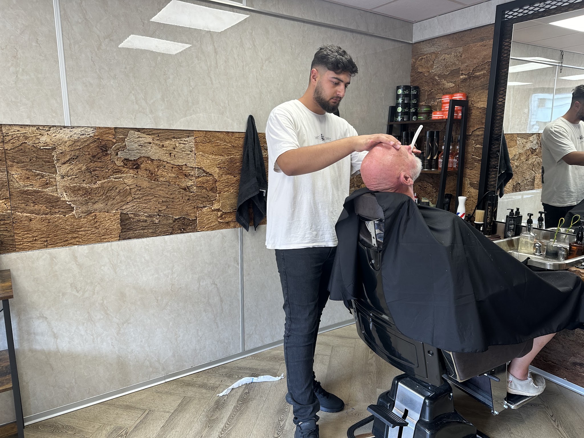 Enes aan het werk bij een klant in de kappersstoel, met de stenen wand en de spiegel erachter