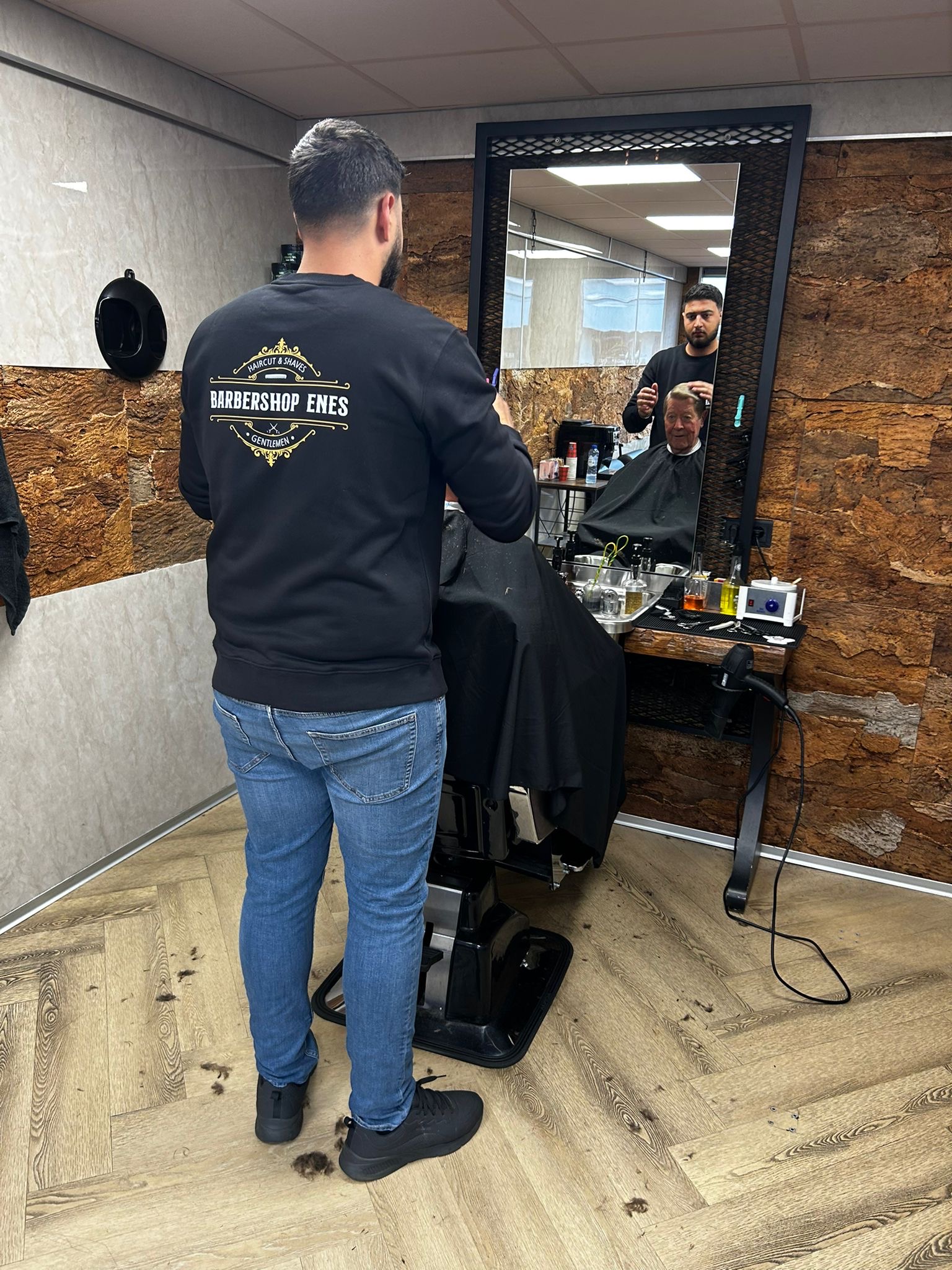 Enes van achteren in een zwarte trui met het Barbershop Enes-logo, aan het werk bij de spiegel